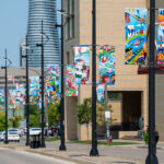 The image shows a row of vibrant street banners designed by Alyah Holmes, displayed along a sidewalk in Mississauga. The colorful illustrations represent different aspects of the community, such as landmarks and local culture.